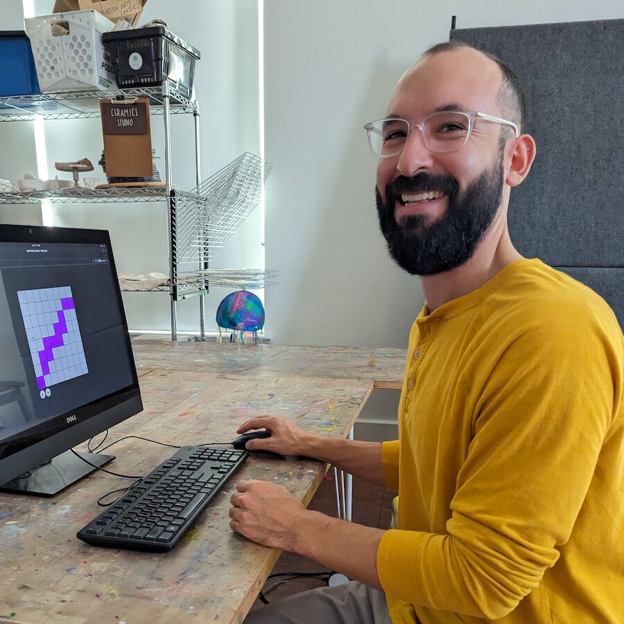 A bearded man in a yellow shirt sitting at a computer while smiling